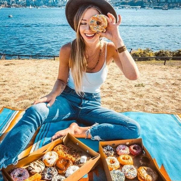 A smiling woman holds a colorful Donut Factory donut over her eye, with boxes of donuts on a blanket by the water.