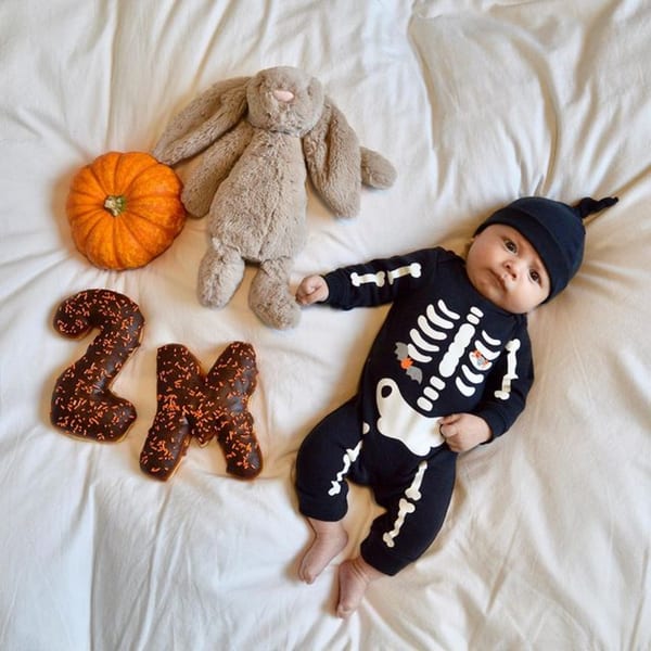 Baby in skeleton costume with a pumpkin, bunny, and "2M" donuts on a white blanket.