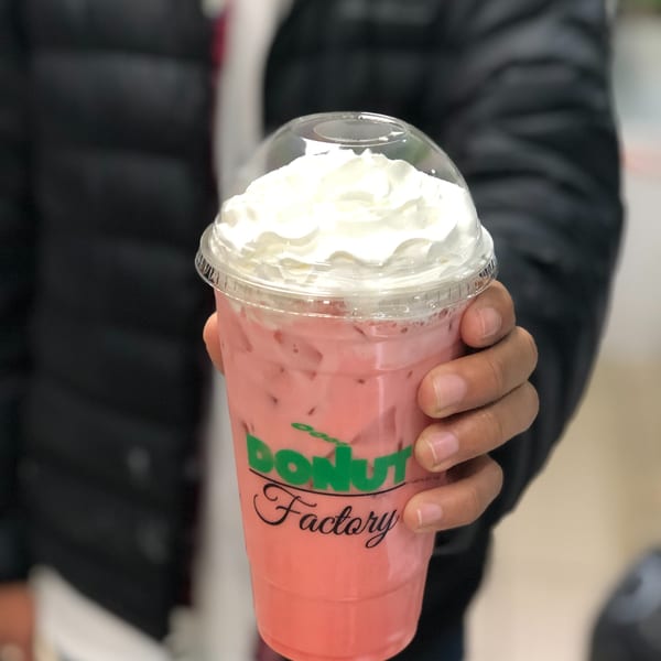 Hand holding a Donut Factory branded cup with a pink drink, ice, and whipped cream.