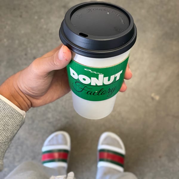 A hand holds a Donut Factory coffee cup, with feet in striped slides visible below.
