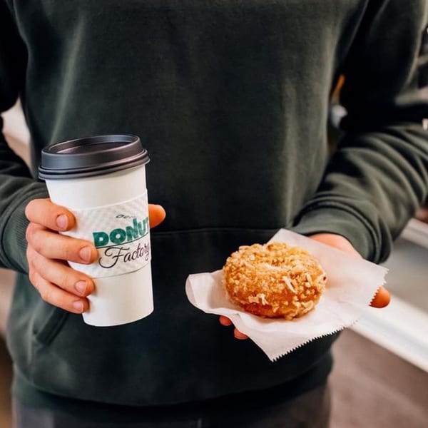 Hands holding a Donut Factory coffee cup and a donut with a crumb topping.