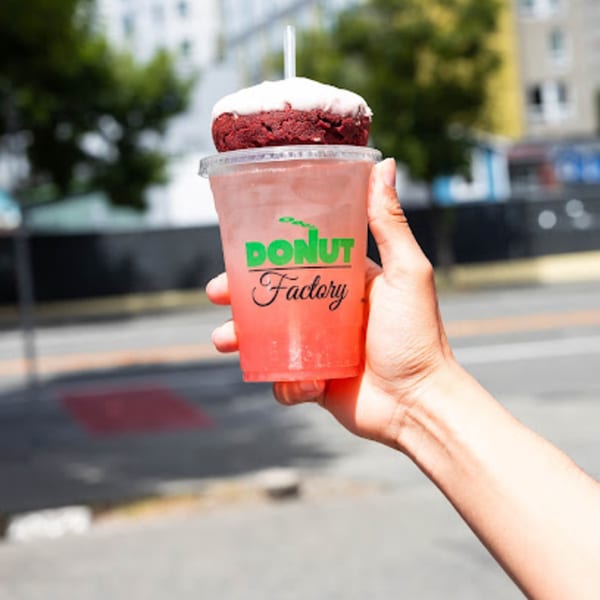 Hand holding a Donut Factory branded cup with a red velvet donut and straw on top.