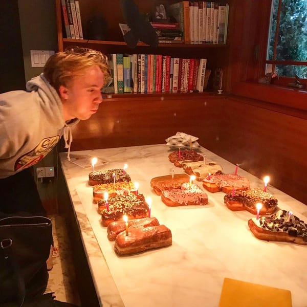 A young man blowing out candles on a table filled with various Donut Factory donuts.