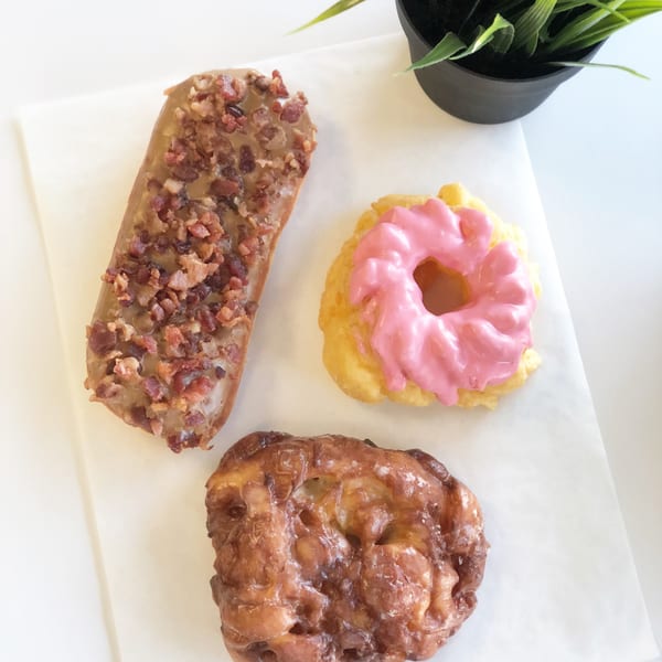 Three Donut Factory donuts: a maple bacon bar, a pink-frosted cruller, and an apple fritter.
