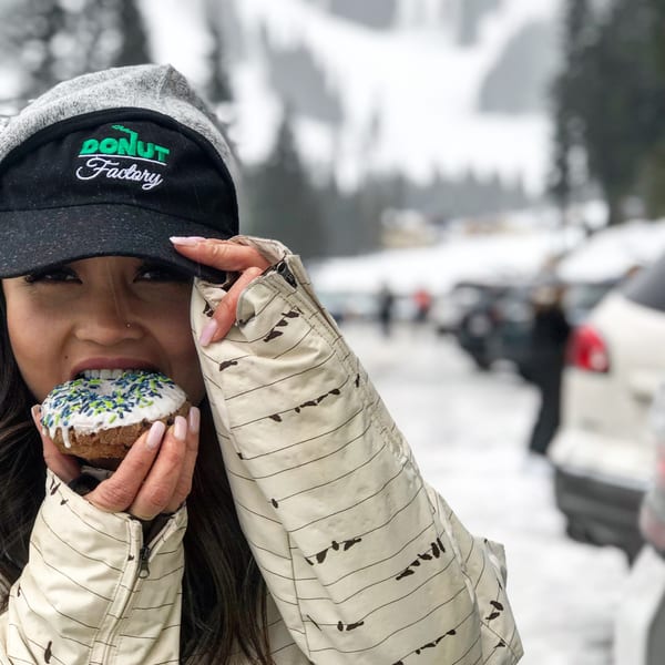 Person wearing a Donut Factory hat taking a bite of a donut with sprinkles in a snowy outdoor setting.