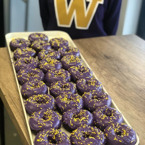 Tray of purple-frosted donuts with yellow sprinkles on a wooden table, with a person in a purple shirt.