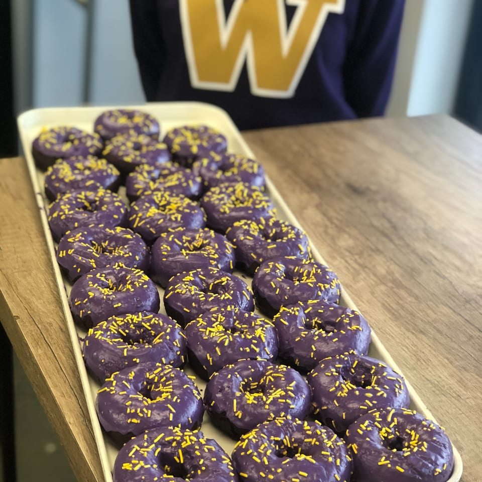 Tray of purple-frosted donuts with yellow sprinkles on a wooden table, with a person in a purple shirt.