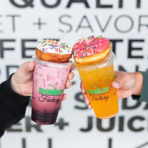 Two hands holding Donut Factory drinks with colorful donuts on top, against a white wall.