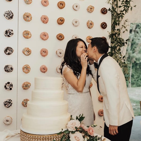 A couple playfully bites a donut from a donut wall next to a wedding cake.