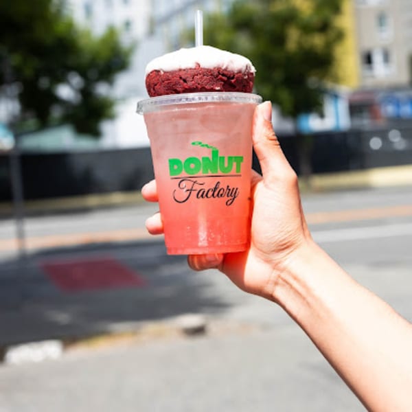 Hand holding a Donut Factory drink with a red velvet donut on top, outdoors.
