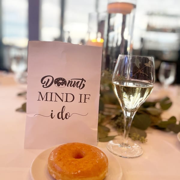 A glazed Donut Factory donut and a glass of champagne next to a "Donut Mind If I Do" sign.