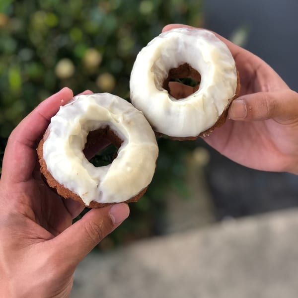 Two hands holding two chocolate donuts with white frosting against a green and gray background.