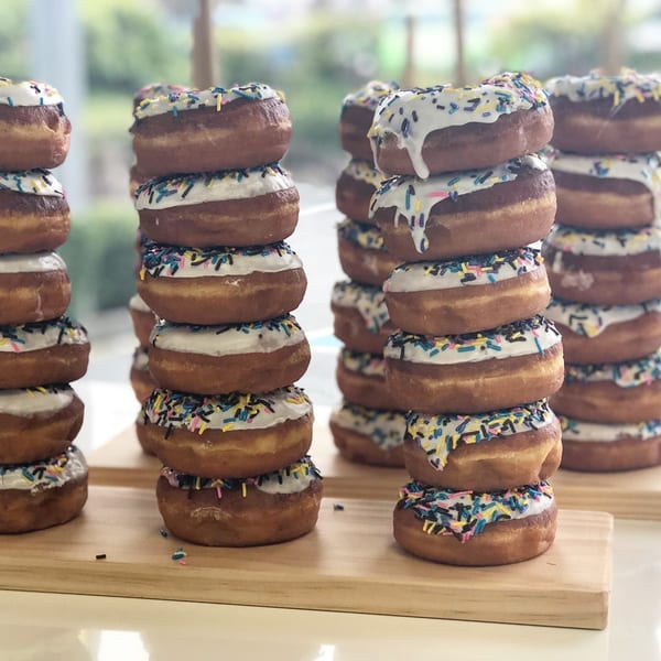 Stacks of Donut Factory donuts with white icing and colorful sprinkles on wooden boards.