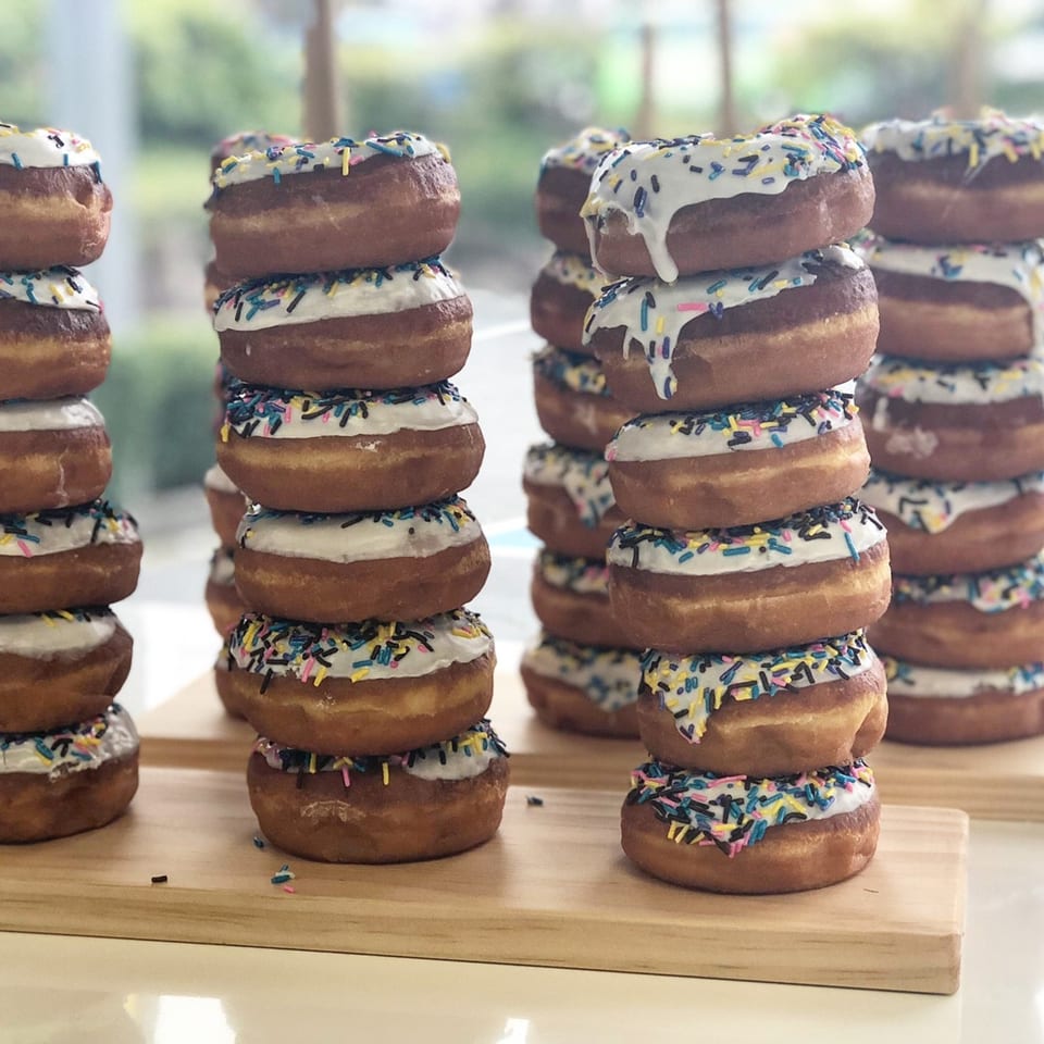 Stacks of Donut Factory donuts with white icing and colorful sprinkles on wooden boards.